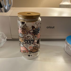 Peut inclure: Un gobelet en verre transparent avec un couvercle en bambou orné d'un motif imprimé d'icônes hivernales, notamment un bonnet rose, des bottes marron, des pommes de pin, des flocons et des tasses de chocolat avec le texte "cozy season".