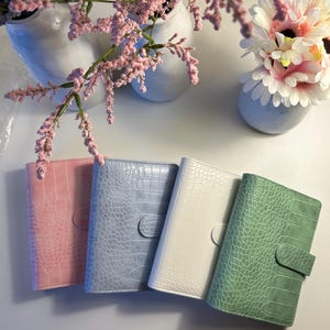 May include: Four rectangular planners with a textured crocodile skin pattern, each featuring a small tab closure. The planners are arranged in a row and come in pastel pink, light blue, white, and sage green colors, each with a matching tab.