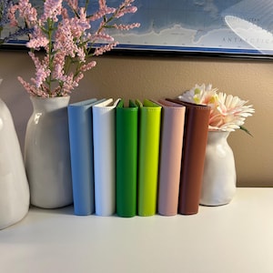 May include: A row of six vertical planners or notebooks standing side by side in a spectrum of colors including light blue, white, grass green, lime green, pale pink, and brown, each featuring a smooth, solid-colored cover and a clean spine.