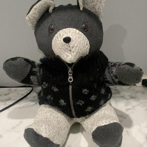 May include: Handmade teddy bear made from socks. The bear has a black head and body, with grey and white accents on the ears, snout, and feet. It wears a black sweater with a zipper and a snowflake pattern. The bear is approximately 25 cm tall.