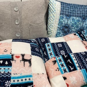 May include: A collection of four square throw cushions. One is a solid light grey knit with three buttons and two pockets. Another is a blue patterned fabric with lace trim. Two feature a patchwork design of pink, white, and navy blue fleece.