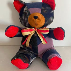 May include: A patchwork teddy bear with a brown nose, red corduroy ears and paws, and a gold and red ribbon bow. The bear's body is made from a mix of patterned fabrics in navy, purple, and black.