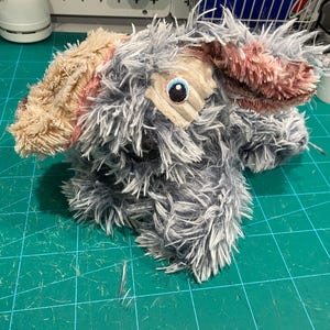 May include: A plush stuffed animal featuring long, shaggy grey and white faux fur. It has large, floppy ears with textured tan and pink fabric patches. One visible eye is embroidered with a blue iris and black pupil on a tan fabric patch.