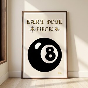 Pode incluir: Uma impressão emoldurada com uma bola oito preta e o número 8 branco. O texto dourado "EARN YOUR LUCK" está acima da bola, com detalhes de estrelas. A impressão está em um fundo creme, com uma moldura de madeira.