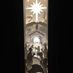 Può includere: Una scena della natività ritagliata in carta bianca con una stella brillante e angeli sopra una mangiatoia con figure dei tre re magi e animali.