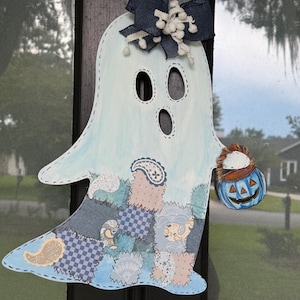 May include: A light blue ghost wall hanging featuring a patchwork-style lower section with various blue, grey, and cream patterns. The ghost has a dark blue fabric bow at the top and holds a small blue jack-o'-lantern bucket with a brown handle.