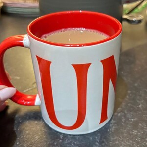 May include: A white ceramic mug with a black handle and the text "But First, Coffee" in a black, handwritten-style font. The mug is on a white surface.