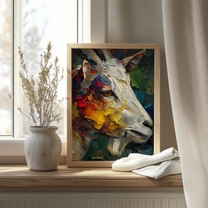 Op de afbeelding: Een ingelijst olieverfschilderij van een geitenkop, weergegeven in een levendige, getextureerde stijl. Het kunstwerk toont een witte geit met een gedetailleerd gezicht, tegen een achtergrond van kleurrijke penseelstreken in tinten rood, geel, groen en blauw. Het schilderij staat op een houten vensterbank.