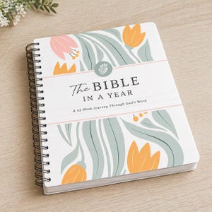 May include: A spiral-bound journal titled "The Bible in a Year" with a floral design. The cover features a white background with illustrations of orange and pink flowers and green leaves. The text "A 52-Week Journey Through God's Word" is also included.