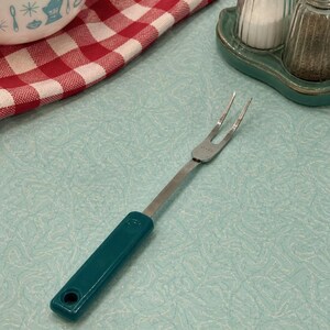 May include: A vintage-style carving fork with a teal handle and stainless steel tines. The fork is designed for serving and is set against a light blue patterned surface, with a red and white checkered cloth in the background.