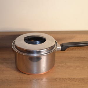 May include: A stainless steel saucepan with a lid and a black handle. The pot has a copper-colored bottom and is sitting on a wooden surface. The lid has a black handle.