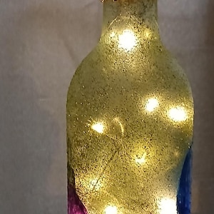 May include: A glass bottle with a frosted, textured yellow surface illuminated by internal string lights. The neck is wrapped in a metallic gold mesh material and topped with a dark brown stopper. Painted circular shapes in purple, blue, and orange are visible.