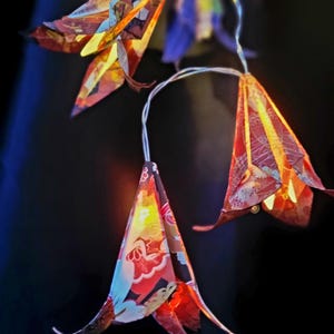 May include: A string of decorative flower-shaped lights. Each flower is made of colorful patterned paper in shades of red, yellow, blue, and orange. The lights are strung on thin, silver wires against a dark background.