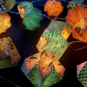 Puede incluir: Una cadena de faroles decorativos de papel origami en tonos naranja, verde y amarillo. Los faroles están iluminados desde dentro, creando un brillo cálido. El papel tiene varios diseños, incluyendo diseños florales y geométricos.