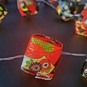 May include: A string of decorative origami paper lanterns in various colors, including red, black, and green. The lanterns are illuminated by small lights, creating a warm glow. The lanterns are arranged on a dark surface.