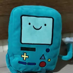 May include: A teal plush toy shaped like a retro video game console character. The toy has a smiling face on the screen, a plus sign, a triangle, and a red circle with a black outline. It has small arms and legs.