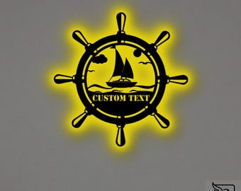 May include: A black metal ship wheel wall sign with a backlit yellow glow. The centre features a silhouette of a sailboat on waves with clouds and a sun in the sky. The words CUSTOM TEXT are printed in black capital letters at the bottom centre.