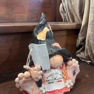 Può includere: Statuetta di gnomo in ceramica con cappello nero e toppa gialla, che tiene un coltello. Lo gnomo ha un grembiule bianco con testo italiano e orlo rosso. Design stravagante.