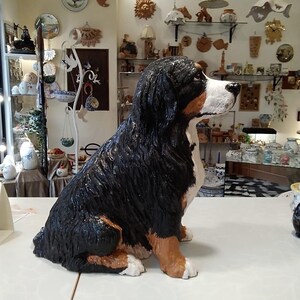 Può includere: Statua di un grande Bovaro del Bernese seduto. Il cane ha pelo nero, bianco e marrone, con una trama dettagliata. La statua è su una superficie bianca, con vari oggetti decorativi sullo sfondo.