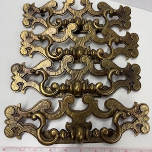 May include: Four antique-style drawer pulls with a gold finish. Each pull features a scrolling design with a central handle and decorative flourishes. The pulls are arranged horizontally. A measuring tape is visible.
