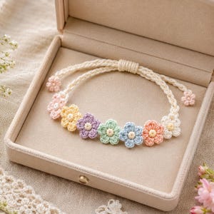 May include: A crochet bracelet featuring colorful flower accents, presented in a beige jewelry box. The bracelet has a cream-colored cord and is adorned with pastel-colored crochet flowers. The box is open, revealing the bracelet's delicate design.
