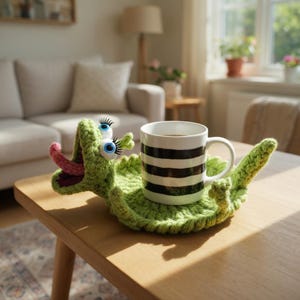 May include: A whimsical green crocheted dragon-shaped coaster with large googly eyes and a pink tongue. A black and white striped mug filled with coffee sits on the coaster, placed on a wooden table. The scene is set in a cozy living room.