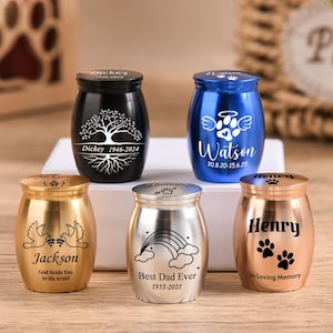 Peut inclure: Plusieurs petits urnes décoratives de différentes couleurs, dont le noir, le bleu, l'or et l'argent. Chaque urne présente des textes et des motifs personnalisés, tels qu'un arbre, des ailes d'ange, un arc-en-ciel et des empreintes de pattes. Ces urnes sont destinées aux funérailles.
