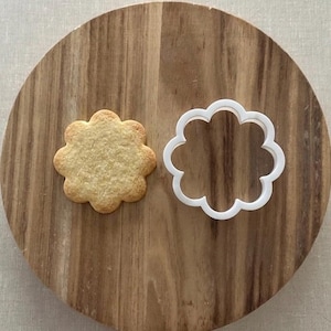 Peut inclure: Un biscuit en forme de fleur, brun doré, à côté d'un emporte-pièce en plastique blanc en forme de fleur. Les deux sont sur une planche à découper ronde en bois. Le biscuit a une surface texturée.