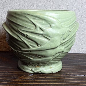 May include: A light green ceramic bowl with a textured, leaf-like design. The bowl has a rounded shape with a small, circular base. The exterior features a detailed, embossed pattern, and the bowl is set on a dark wooden surface.