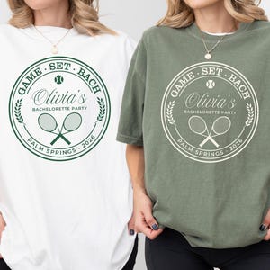 May include: Two t-shirts, one white and one olive green, featuring a circular design with tennis rackets and the text "GAME SET BACH Olivia's BACHELORETTE PARTY PALM SPRINGS 2026". The design is in green and white.