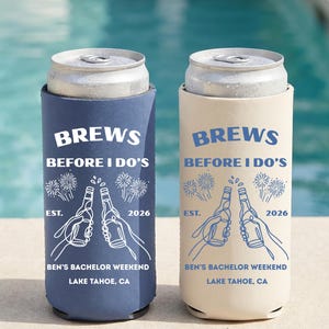 Puede incluir: Dos enfriadores de latas delgados, uno azul marino y otro crema, cada uno con dibujos lineales en blanco y azul de dos manos brindando con botellas de cerveza, fuegos artificiales y el texto: BREWS BEFORE I DO'S, EST. 2026, BEN'S BACHELOR WEEKEND, LAKE TAHOE, CA.
