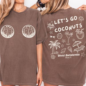 May include: Brown t-shirt with a white coconut design on the front. The back of the shirt features the text "LET'S GO COCONUTS" with beach-themed illustrations, including a palm tree, cocktail, and sunglasses. "Olivia's Bachelorette Miami, FL" is also printed on the back.