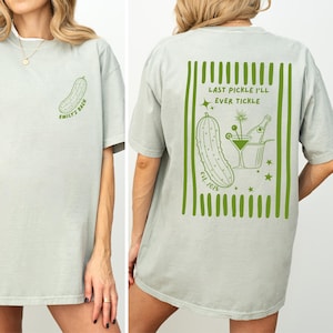 May include: A light sage green t-shirt featuring a small pickle graphic on the front chest with the text EMILY'S BACH. The back displays a large green striped design with a pickle, a martini glass, a champagne bottle, and the text LAST PICKLE I'LL EVER TICKLE EST. 2026.