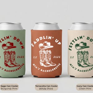 Puede incluir: Tres enfriadores de latas en salvia, terracota e marfil. Cada uno presenta un sombrero de vaquero y botas con el texto "Settlin' Down" o "Saddlin' Up", "Est 2026" y "Mia's Bachelorette". El enfriador de salvia tiene una impresi&oacute;n burdeos, el terracota tiene una impresi&oacute;n blanca y el marfil tiene una impresi&oacute;n verde.