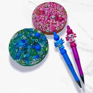 May include: A collection of handmade axolotl-themed items. Includes two pens, one blue and one purple, each topped with an axolotl figure. Also, two circular resin coasters, one pink and one blue, with axolotl and bead details.