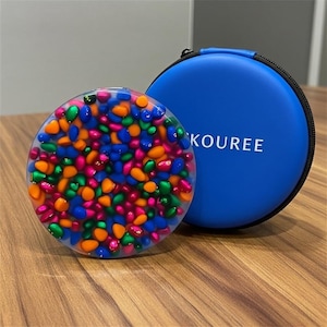 Puede incluir: Un posavasos redondo y transparente lleno de cuentas de colores en forma de guijarros en tonos naranja, rosa, verde y azul. Una funda redonda azul con la palabra 'KOUREE' en blanco está junto al posavasos. El posavasos y la funda están sobre una superficie de madera.