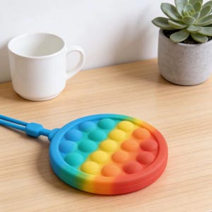 May include: A circular silicone sensory toy featuring a rainbow gradient of blue, yellow, orange, and red colors with multiple rows of pushable bubbles. The toy includes a thin blue strap attached to the side for carrying or hanging purposes.