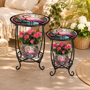 May include: Two black metal plant stands with round tops and lower shelves. Each features a stained glass-style tabletop and matching planter pot, both decorated with a pink flamingo illustration set against a blue and green water background.