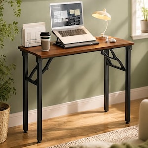 May include: A rectangular folding desk with a dark wood grain surface and black metal legs. The desk features a hinged support mechanism on the sides. On the desk sits a laptop on a stand, a white desk lamp, a paper organizer, and a coffee cup.