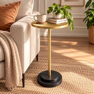 May include: A round side table with a gold-colored metal top featuring a radial sunburst texture, a thin gold-colored central post, and a heavy, circular black metal base. The table surface holds a speckled ceramic mug and a small potted plant.