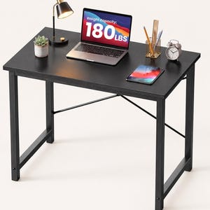 May include: A black rectangular desk with a metal frame and X-shaped support bars. On the surface sits a laptop displaying the text Weight capacity: 180 LBS, a small potted succulent, a black desk lamp, a gold wire pencil holder, and a tablet.