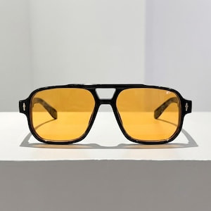 May include: Black square-framed sunglasses with orange-tinted lenses. The sunglasses have a double bridge and a thick frame. The temples feature a decorative element. The sunglasses are sitting on a white surface.