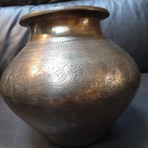 May include: Antique brass pot with intricate etched designs. The pot has a rounded body, a wide base, and a slightly flared rim. The surface is covered in detailed patterns, including floral and bird motifs. The pot's aged appearance suggests it is a vintage item.