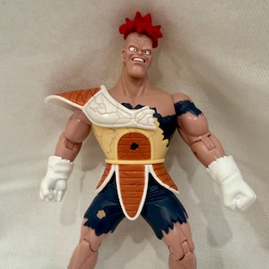 May include: A detailed action figure of a muscular character with red spiky hair. The figure wears torn blue shorts, white gloves, and boots with blue stripes. The character's armor is tan and brown, with a cracked chest plate.