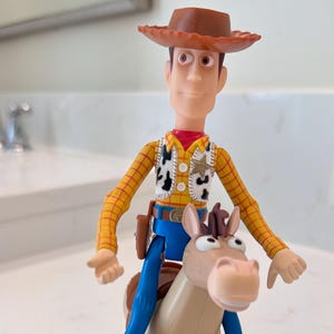 May include: A Woody action figure from the Toy Story franchise, wearing a brown cowboy hat, yellow plaid shirt, vest with cow print, blue trousers, and brown boots. The figure is riding a tan horse.