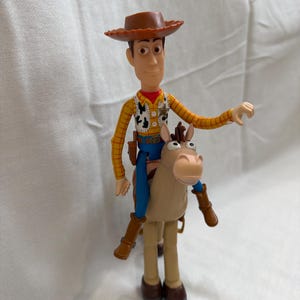 May include: A Woody action figure riding a tan horse. Woody wears a brown cowboy hat, a yellow plaid shirt, a vest, blue trousers, and brown boots. The horse has a tan body, brown legs, and a light brown face.