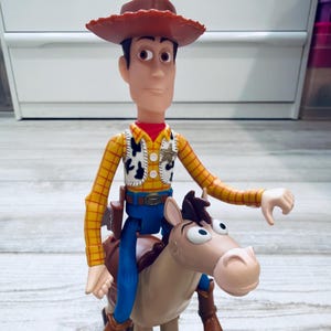 May include: A Woody action figure from the Toy Story franchise, wearing a cowboy hat, yellow plaid shirt, vest, blue trousers, and brown boots. The figure is riding a tan horse with a brown saddle.