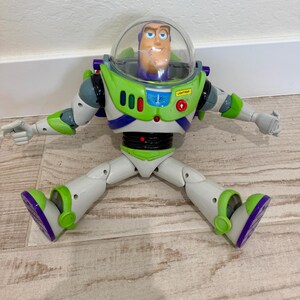 May include: A Buzz Lightyear action figure from the Toy Story franchise. The toy is primarily white, green, and purple, with a clear plastic helmet. The figure has articulated limbs and is posed with arms and legs spread.