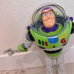 May include: A Buzz Lightyear action figure in a classic green, white, and purple spacesuit. The toy has a clear helmet, a smiling face, and the word "LIGHTYEAR" on the chest. The figure is pointing with its left arm.