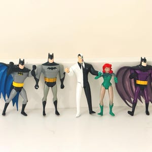May include: A set of five action figures featuring two Batman figures with blue and purple capes, a grey Batman figure, a Two-Face figure in a split black and white suit, and a Poison Ivy figure in a green outfit with red hair.